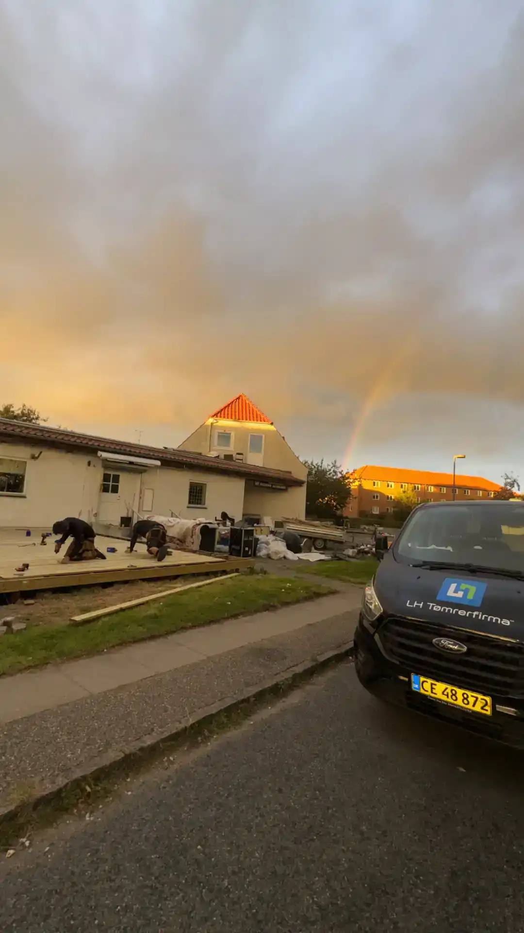 - Mindst 100 tegn: Tømrerfirma arbejder på byggeri under dramatisk himmel med regnbue i baggrunden.