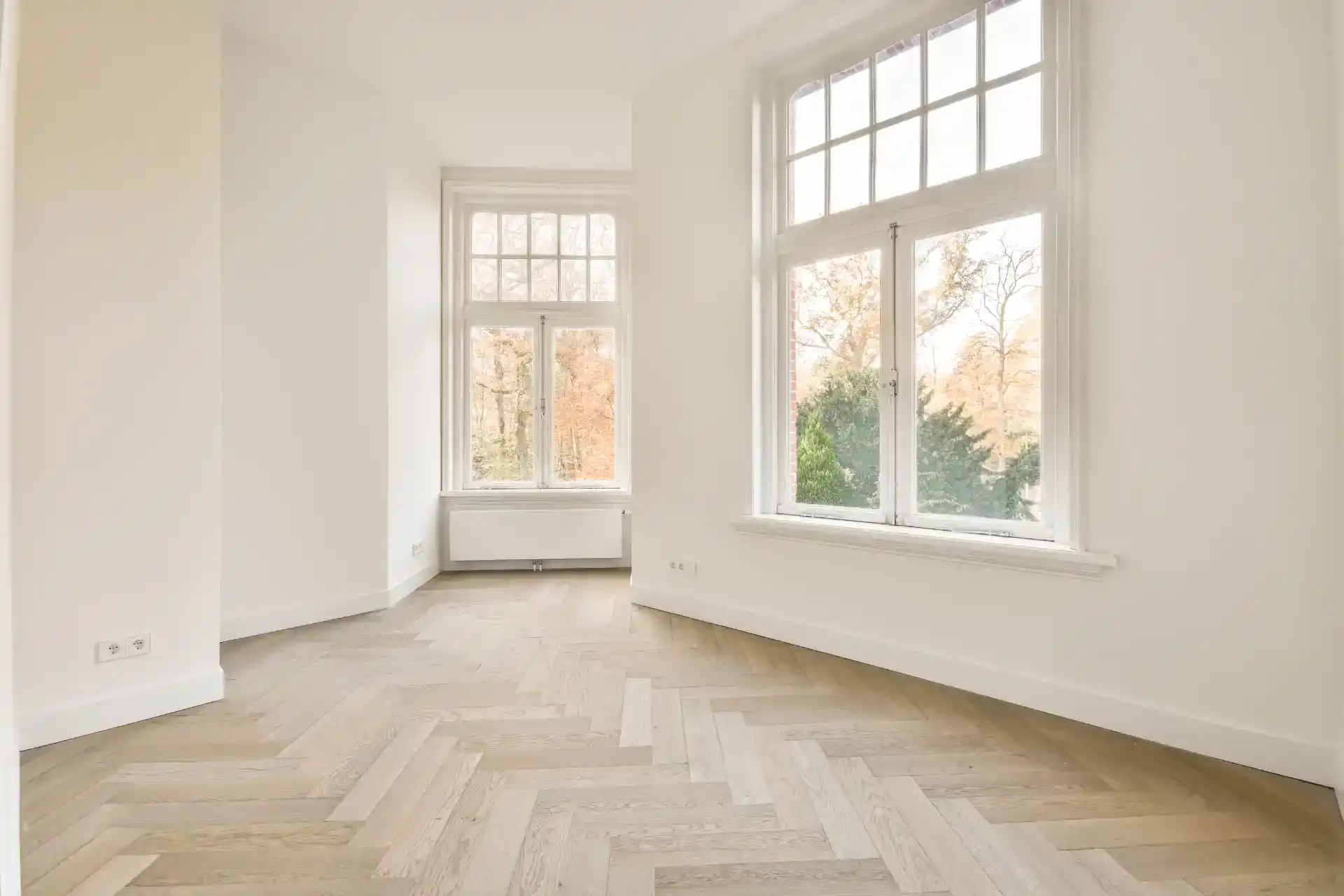 Hvide, store vinduer i rummelig, lyse stue med træparketgulv og udsigt til natur.